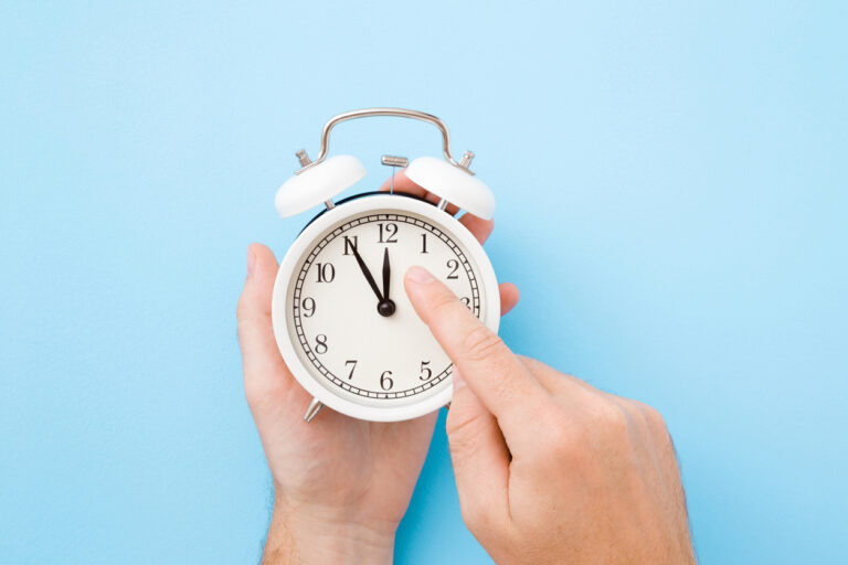 Man hand holding white alarm clock. Finger pointing to arrow of twelve o'clock. Light pastel blue background. Time change concept. Closeup. Top view.